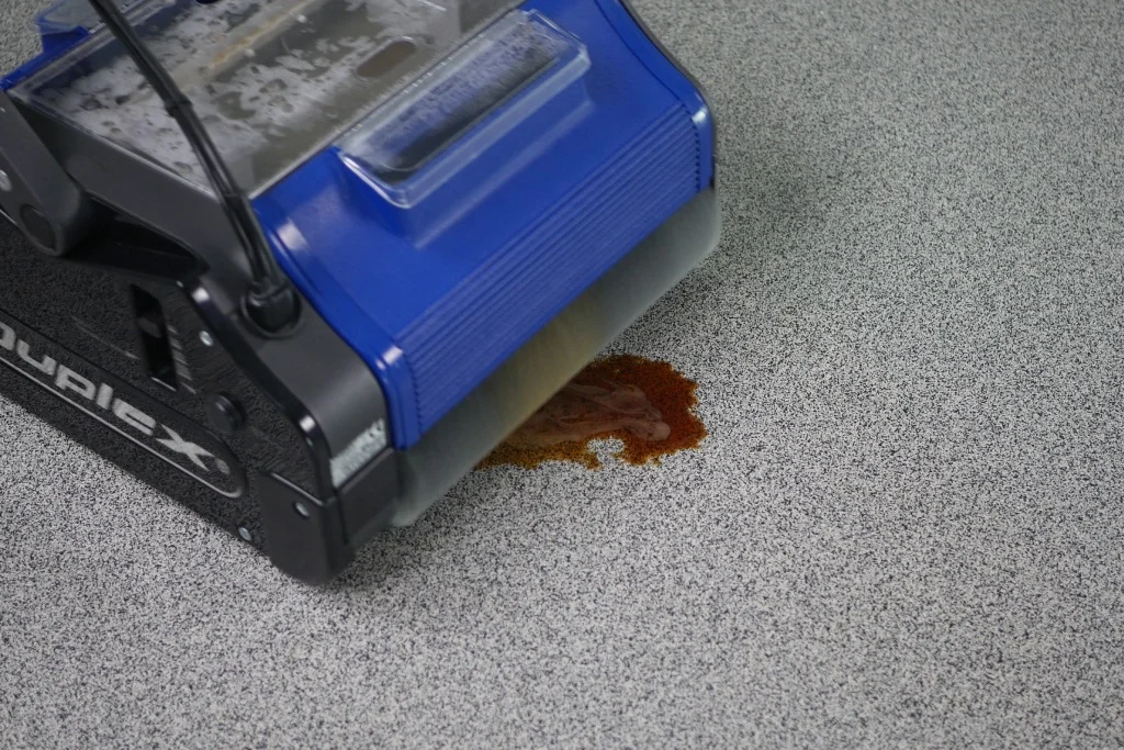 Compact commercial floor cleaning machine lifting a red stain from a speckled vinyl floor.
